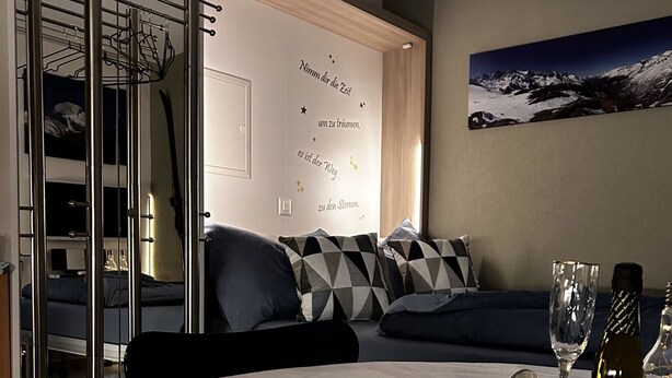 A modern hotel room with a bed, a dining table with a magazine, a wine glass, and a bottle. There is a view of mountains on the wall. - Evelyne by Interhome (Saas-Fee)