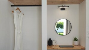 Une pièce dans laquelle on peut voir une robe blanche suspendue, un miroir rond reflétant un arbre, une étagère en bois avec un chapeau, un ordinateur portable, un vase et une plante en pot.