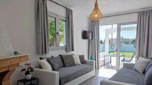 Un salon moderne avec un canapé, une petite table sur laquelle est posée une plante, un téléviseur fixé au mur et une vue sur l’extérieur grâce à de grandes fenêtres.