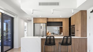 A modern kitchen with a marble island, wooden cabinets, and two black bar stools. - The Monterey Apartments by Urban Rest (Kangaroo Point)