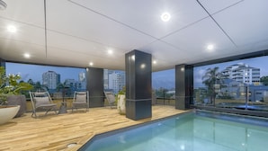 A modern indoor pool area with a wooden floor, two lounge chairs, and a large window offering a city view. - The Monterey Apartments by Urban Rest (Kangaroo Point)