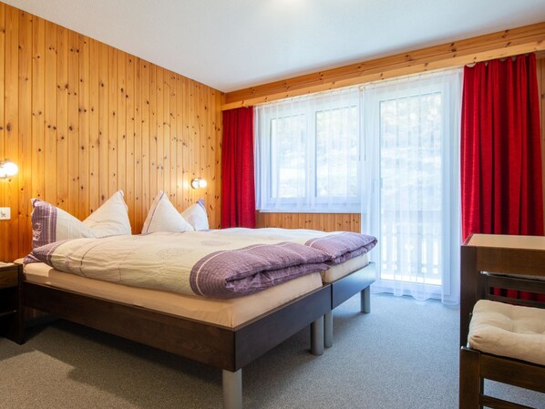 A wooden-paneled room with a bed, a nightstand, a chair, and a window with red curtains.