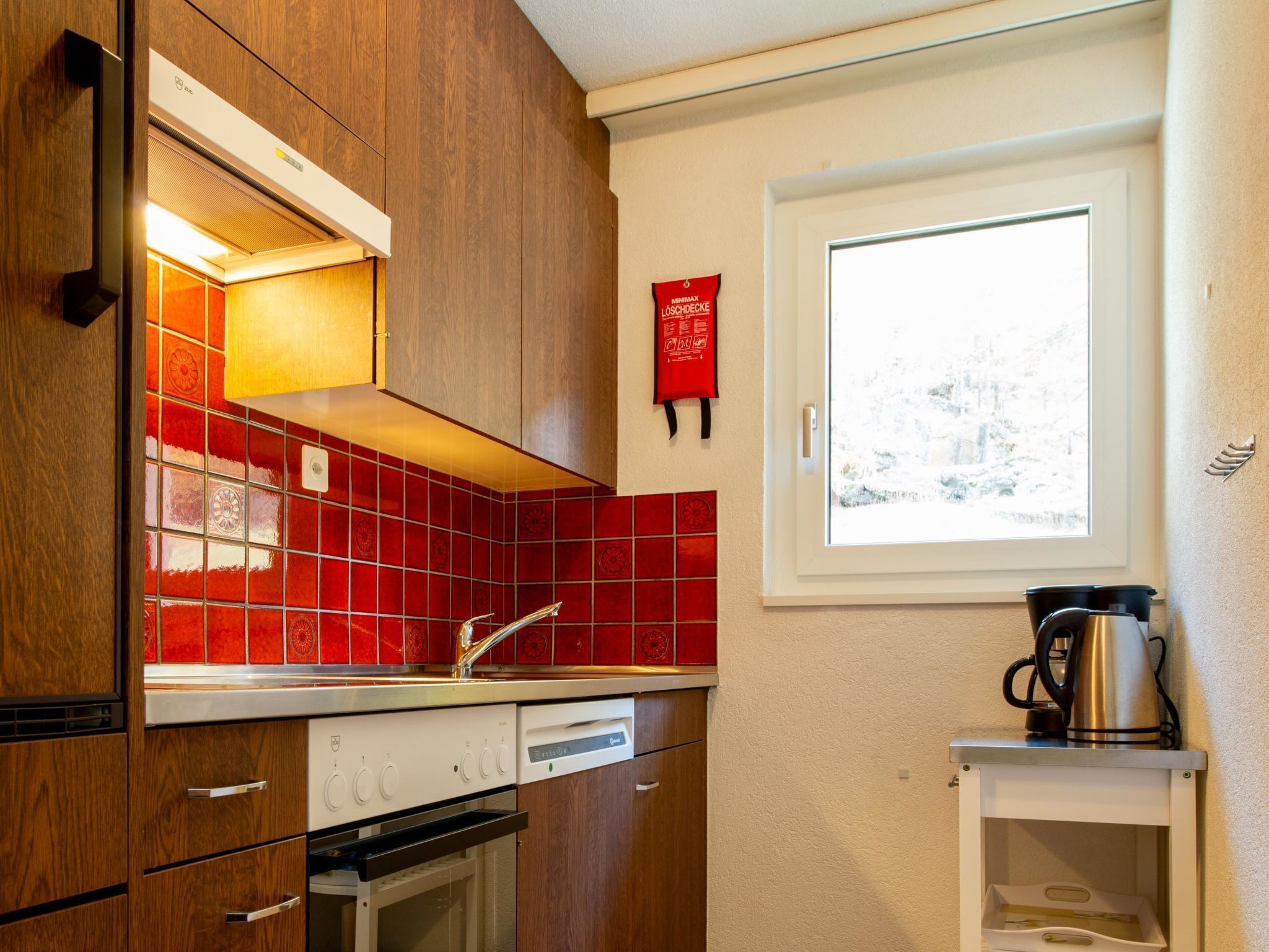 Una cocina moderna con armarios de madera, baldosas rojas, horno empotrado y ventana con vistas a un paisaje nevado.