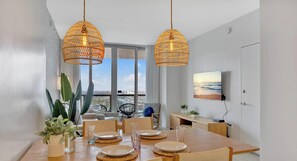 Zona pranzo con tavolo in legno apparecchiato per quattro persone, due lampade pendenti in vimini, un'ampia finestra con vista e una TV a schermo piatto montata a parete.