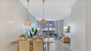 Et moderne spiseområde med højt til loftet, hængende lamper, et spisebord dækket til fire personer og udsigt til solnedgangen gennem store vinduer.