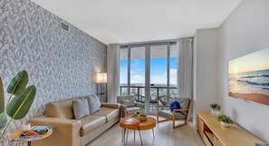 Un soggiorno moderno con divano, poltrona e tavolino da caffè, ampie finestre con vista sulla città e un quadro raffigurante l'oceano.