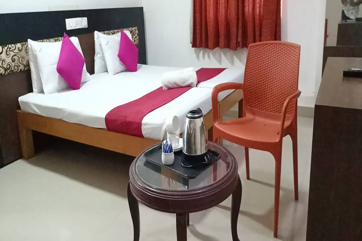 Una cama con una colcha roja, dos almohadas, una mesita con una tetera y una taza, y una silla roja.