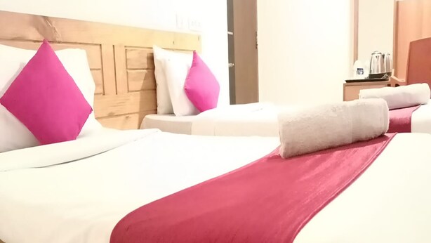 A hotel room with two beds, each with white bedding and pink pillows. There is a wooden headboard, a nightstand with a kettle, and a door in the background. - Aayush Residency (Chennai)