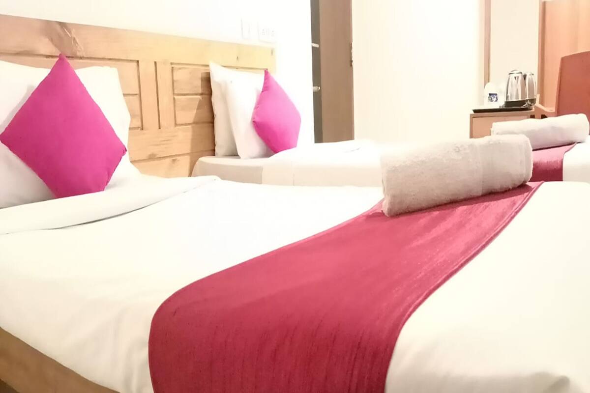 Habitación de hotel con dos camas, cada una con ropa blanca y almohadas rosas. Se observa un cabecero de madera, una mesita de noche con una tetera y una puerta al fondo.
