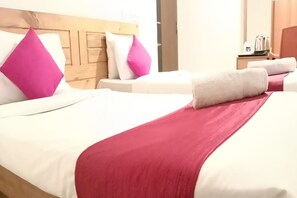Habitación de hotel con dos camas, cada una con ropa blanca y almohadas rosas. Se observa un cabecero de madera, una mesita de noche con una tetera y una puerta al fondo.