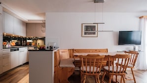 A modern kitchen with wooden cabinets, a dining area with a wooden table and chairs, a TV mounted on the wall, and a ceiling light fixture. - Haus Sunneschy Whg. 202 by Interhome (Parpan)