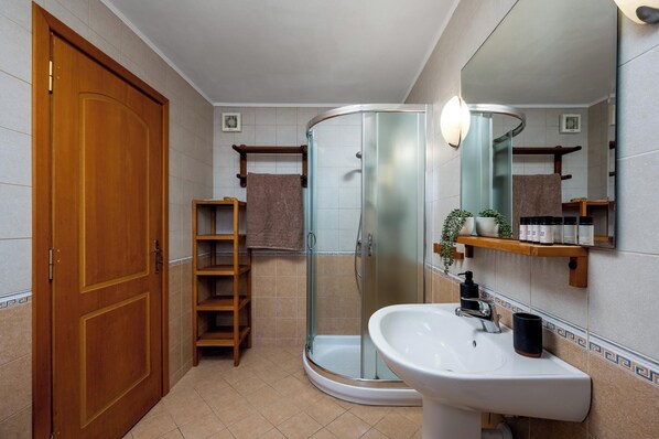 Un baño con puerta de madera, cabina de ducha con vidrio, lavamanos blanco y estante con artículos de aseo.