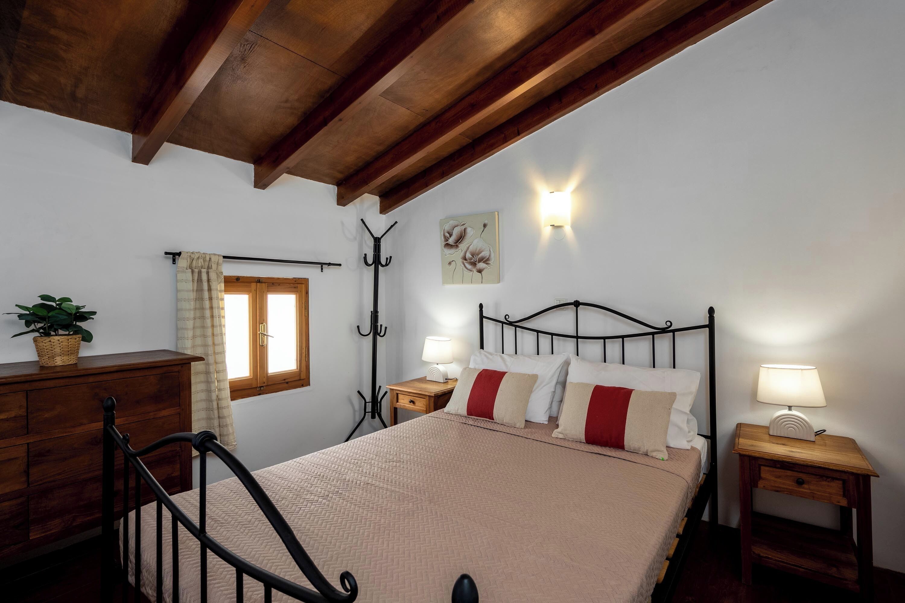 A bedroom with a wooden ceiling, a bed with a beige quilt, two bedside tables with lamps, a wooden dresser, a window with curtains, and a wall-mounted light fixture.