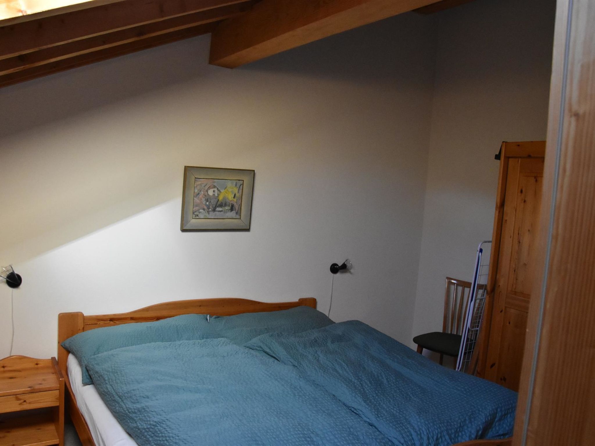 A bedroom with a wooden bed, a blue blanket, a framed picture on the wall, and a wooden door.