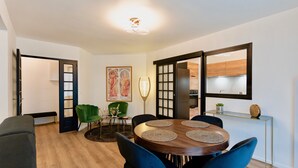 A modern dining area with a round wooden table, blue chairs, and a green chair. There is a large mirror on the wall and a painting above the table. - Résidence Champenoise Lancelot Rouyer (Épernay)