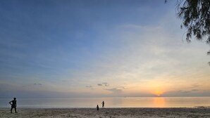 Uma praia com um pôr do sol ao fundo, apresentando uma faixa de areia e algumas pessoas em pé.