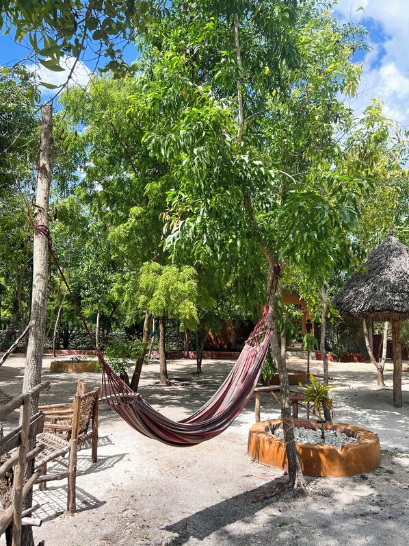 A hammock suspended between trees in a garden area with a thatched-roof structure nearby.