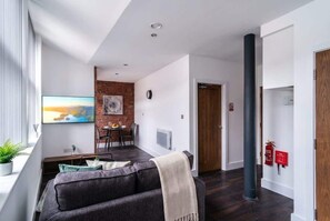 Una sala moderna con un sofá oscuro, una zona de comedor con una pared de ladrillo decorativa y un ventanal grande.
