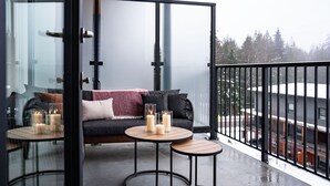 A balcony with a glass partition, a sofa, two small tables with candles, and a view of a snowy landscape. - Epic Mountain Views & AC | 2.5km to RMR & Town⁠ (Revelstoke)