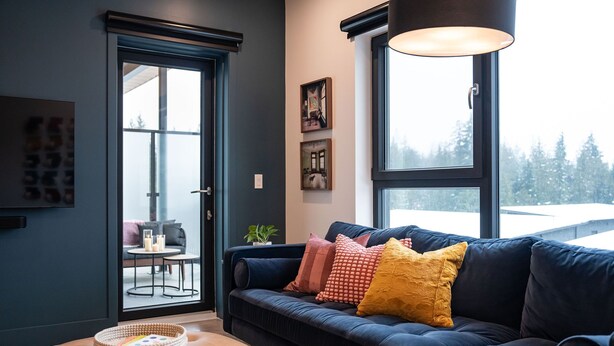 A modern living room with a dark blue sofa, a colorful throw blanket, and a wooden basket on a leather ottoman. - Epic Mountain Views & AC | 2.5km to RMR & Town⁠ (Revelstoke)