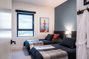 A bedroom with two beds, a window with a view of a snowy landscape, a framed picture of a red tree, and a bedside lamp. - Epic Mountain Views & AC | 2.5km to RMR & Town⁠ (Revelstoke)