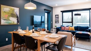 A modern dining area with a wooden table set for a meal, a blue sofa, and a large window with a view of trees. - Epic Mountain Views & AC | 2.5km to RMR & Town⁠ (Revelstoke)