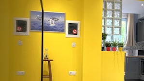 Een helder gele kamer met een keukenhoekje, een ladder en een schilderij van dolfijnen.