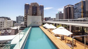 A rooftop pool with wooden decking and white umbrellas, overlooking a cityscape with various buildings. - Studio With City Views (Cape Town)