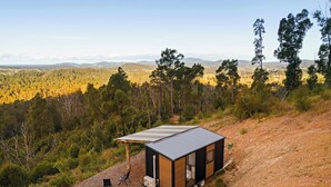 Une petite cabane moderne, dotée d’un toit en métal, située sur un chemin de terre, au milieu d’une forêt dense et de collines.