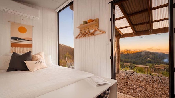 Une chambre à coucher avec un lit, des oreillers, une étagère fixée au mur et une grande fenêtre donnant sur un paysage pittoresque.