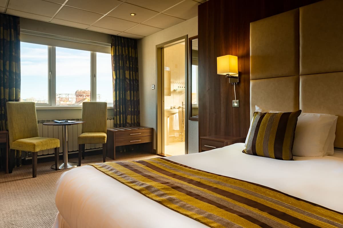 A hotel room with a large bed, a desk with two chairs, a window with curtains, and a bathroom visible through an open door.