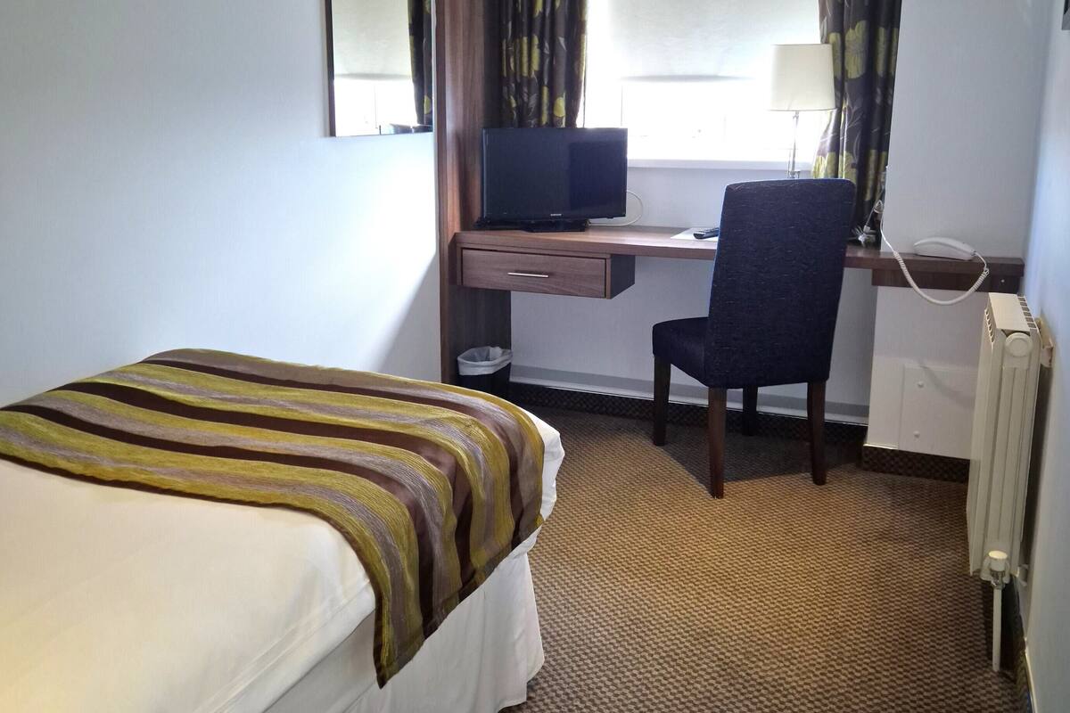 A hotel room with a bed, a desk with a computer, a chair, a telephone, and a window with curtains.