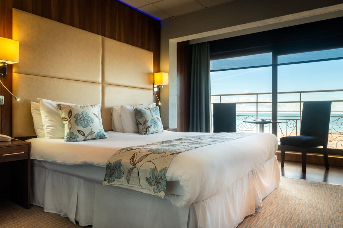 A hotel room with a large bed, two bedside lamps, a view of the sea through large windows, and a balcony with a chair.
