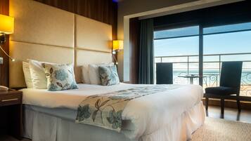 A hotel room with a large bed, two bedside lamps, a view of the sea through large windows, and a balcony with a chair.