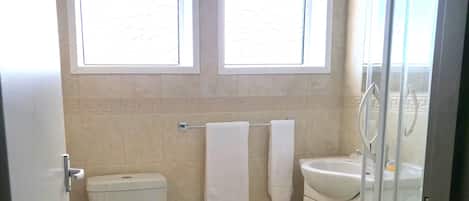 A bathroom with a toilet, sink, and shower. There are two windows above the toilet and a towel rack with towels.