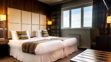 A hotel room with a large bed, two bedside tables, a chair, a window with curtains, and a radiator.
