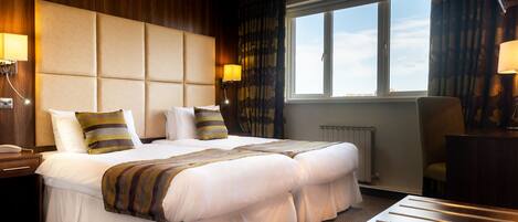 A hotel room with a large bed, two bedside tables, a chair, a window with curtains, and a radiator.