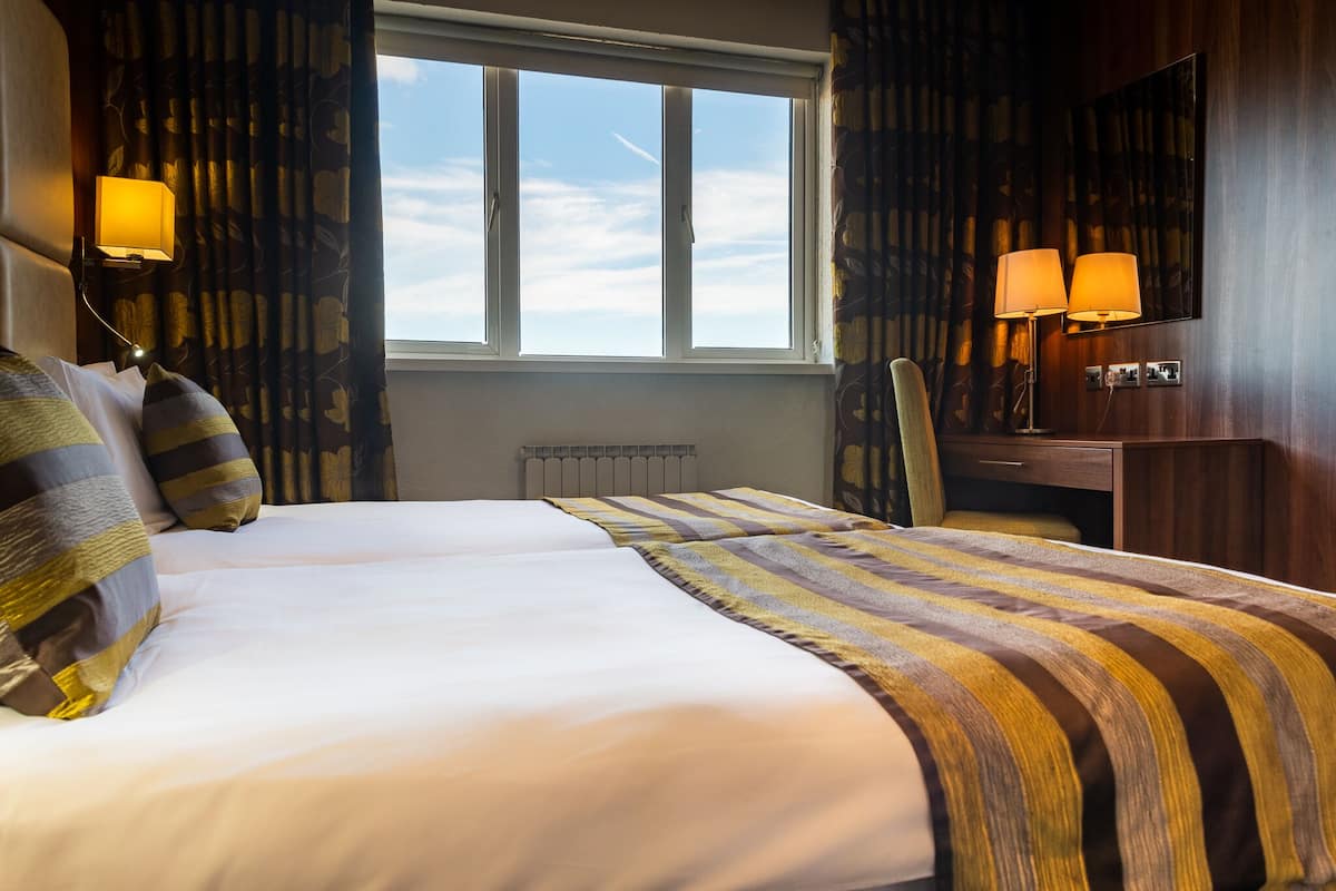 A hotel room with two beds, a large window, a desk with a chair, and two wall-mounted lamps.