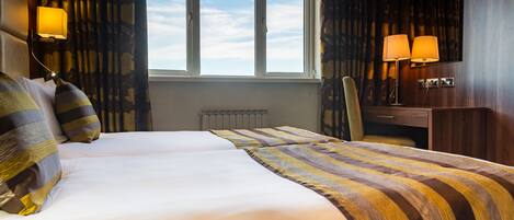 Une chambre d’hôtel avec deux lits, une grande fenêtre, un bureau avec une chaise et deux lampes fixées au mur.
