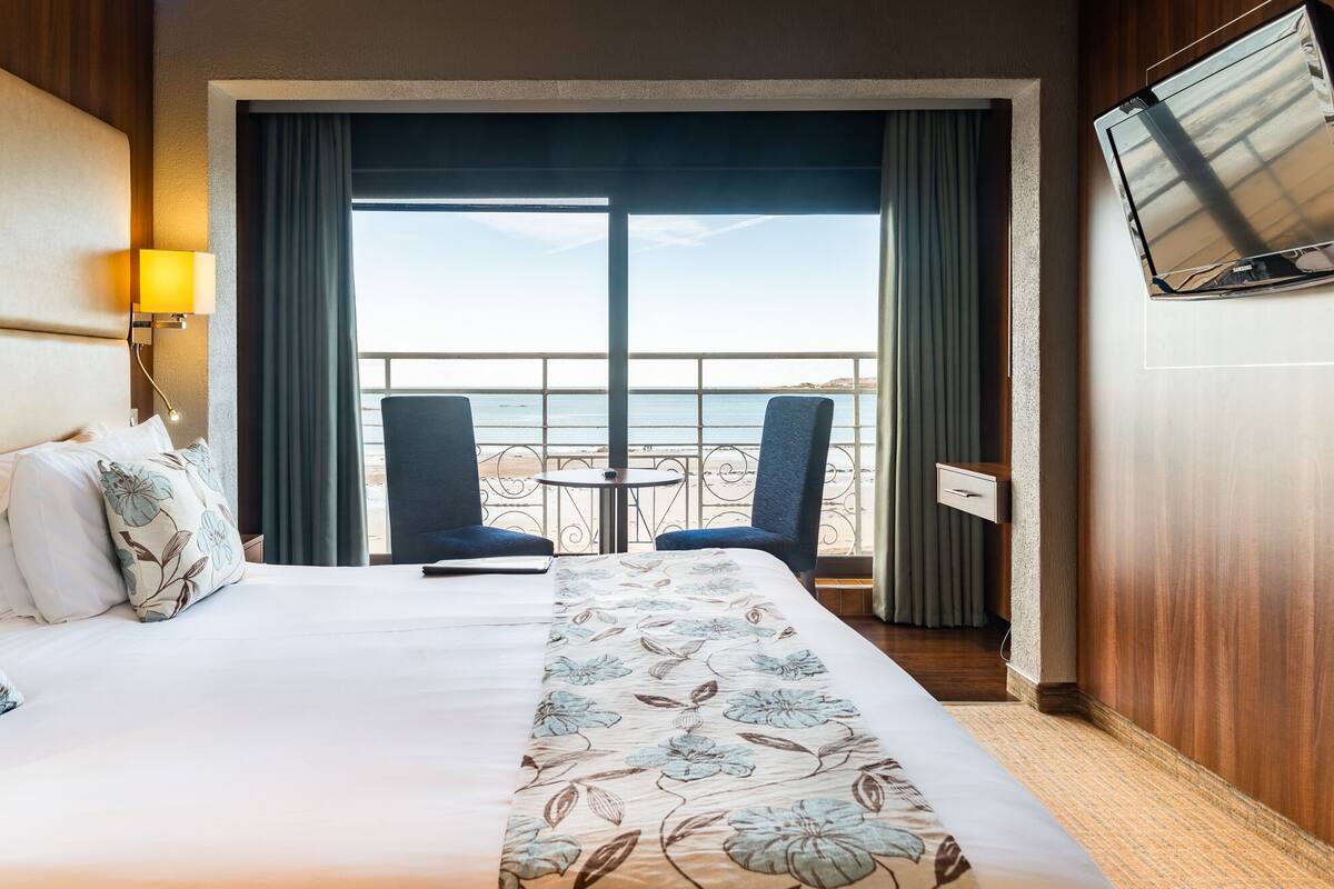 A hotel room with a bed, two chairs, a small table, a TV, and a view of the sea.
