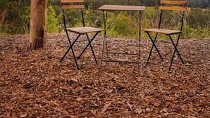 Ulkona sijaitseva oleskelualue, jossa on kaksi puista tuolia ja pöytä. Alue on aaltopeltikatoksen alla, ja sieltä avautuu näkymä metsäiseen maisemaan.