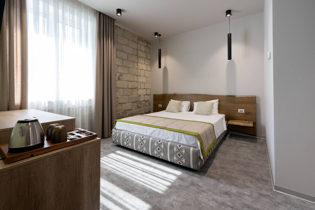 A hotel room with a bed, a wooden desk with a kettle, a brick wall, and a window with curtains.
