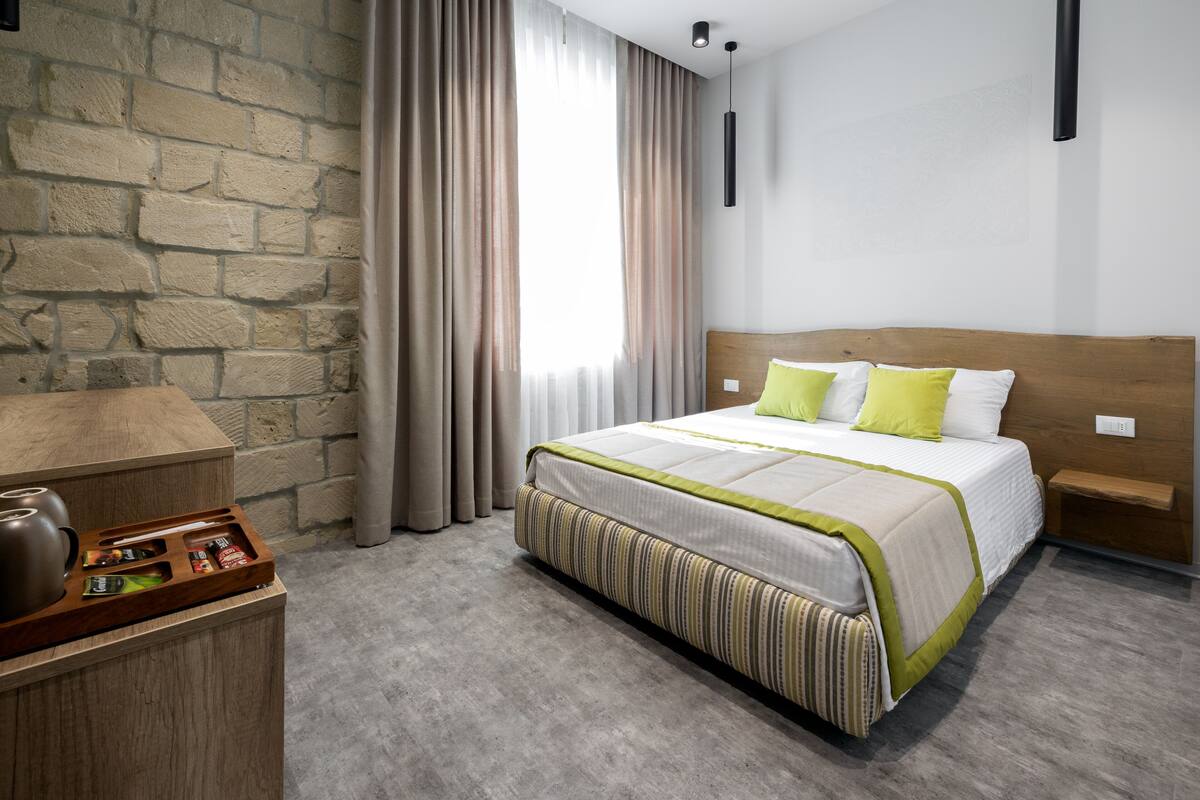 A hotel room with a stone wall, a wooden headboard, a bed with a striped bedspread, and a bedside table with a lamp.