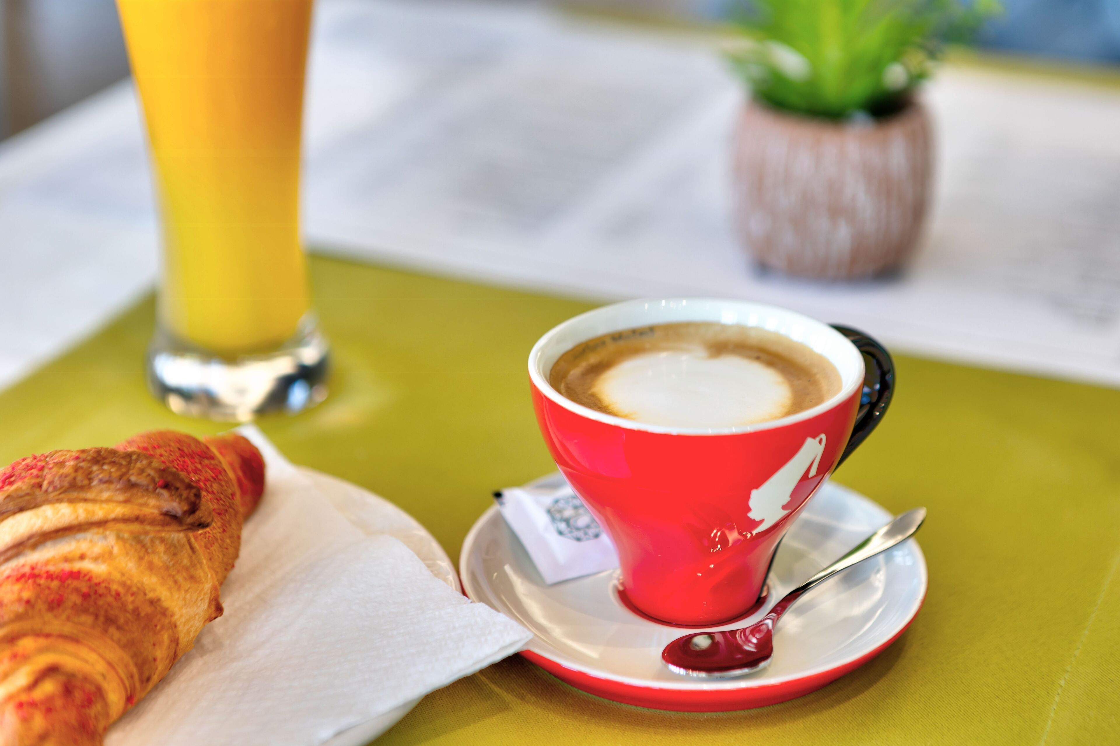 A breakfast setting with a croissant, a red cup of coffee, and a glass of orange juice on a table.