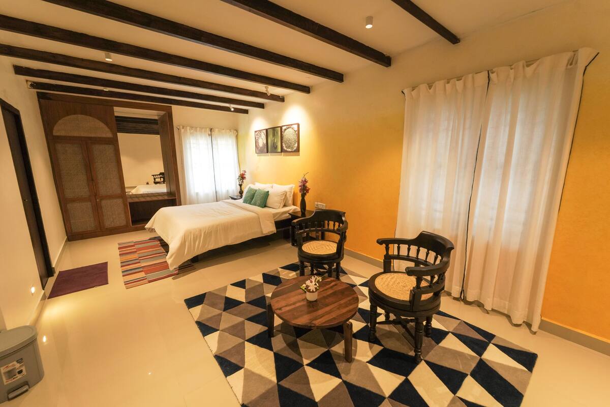 Quarto de hotel com cama, duas cadeiras, uma mesa pequena, tapete e janela com cortinas.