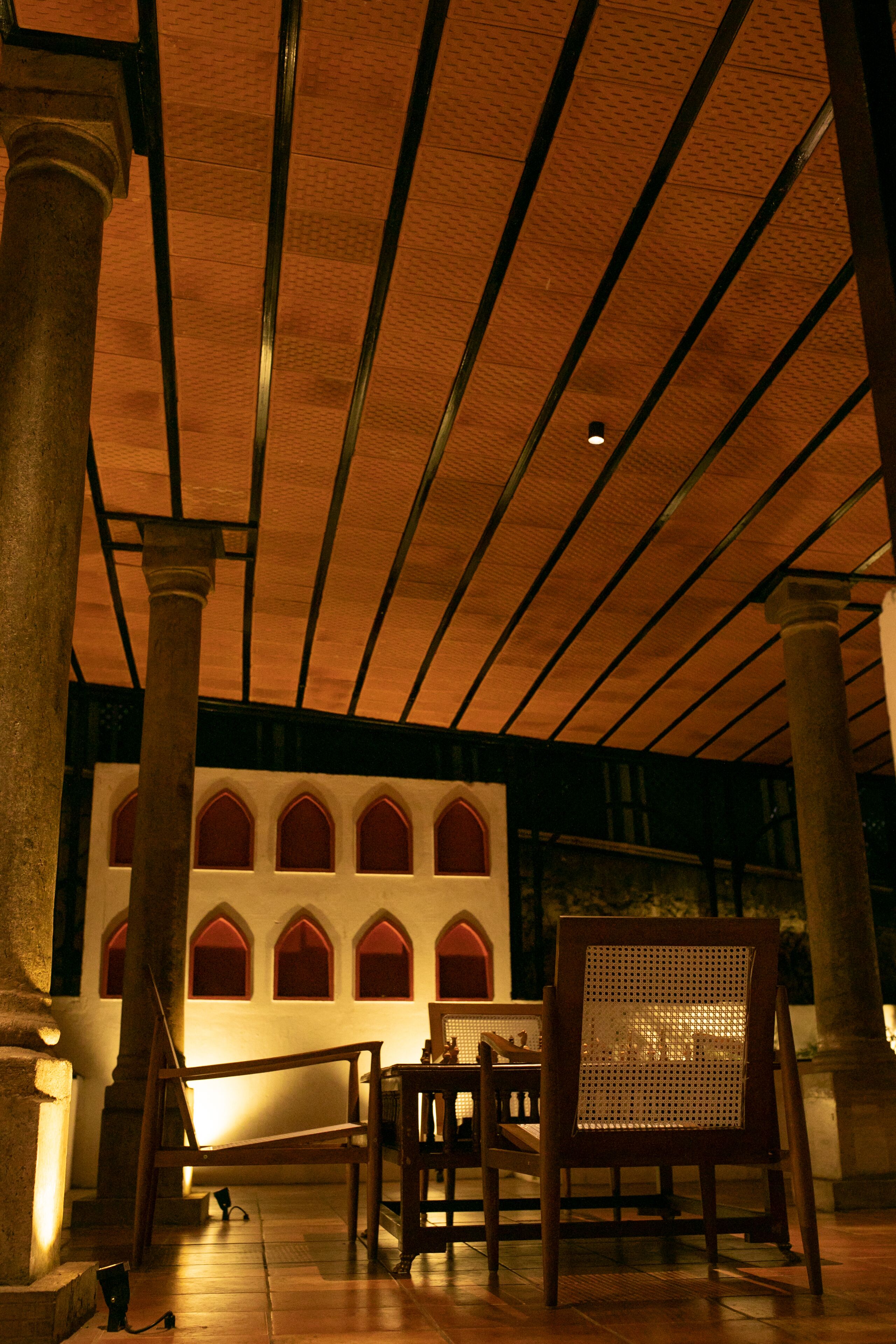 A room with a high ceiling, wooden beams, and a series of arched windows. There are wooden chairs and a table in the foreground.