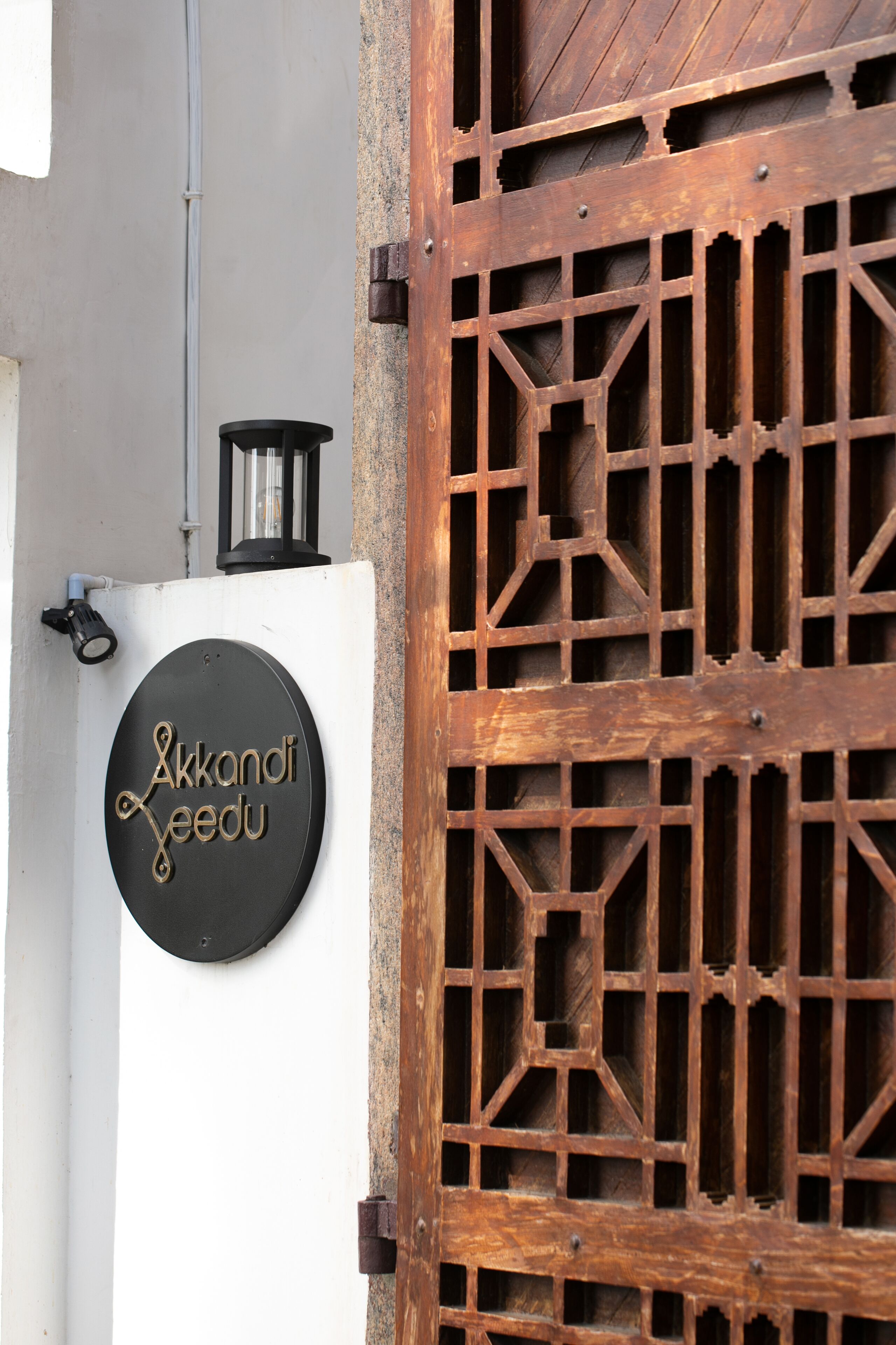 A wooden gate with a geometric pattern, a black circular sign with gold lettering, and a small black lantern on top.