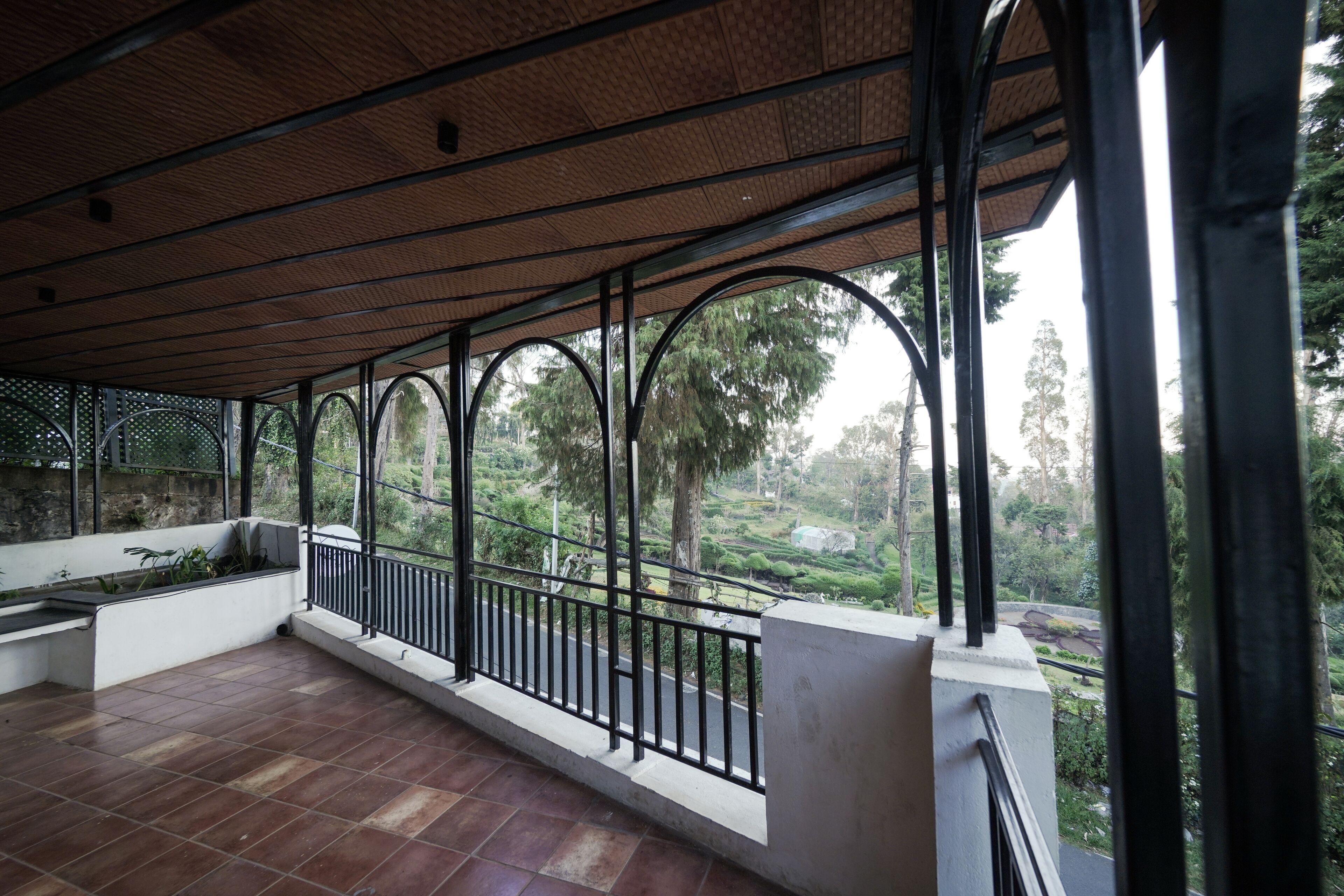 Uma varanda coberta com piso de azulejos, grades de metal e vista para um jardim paisagístico.