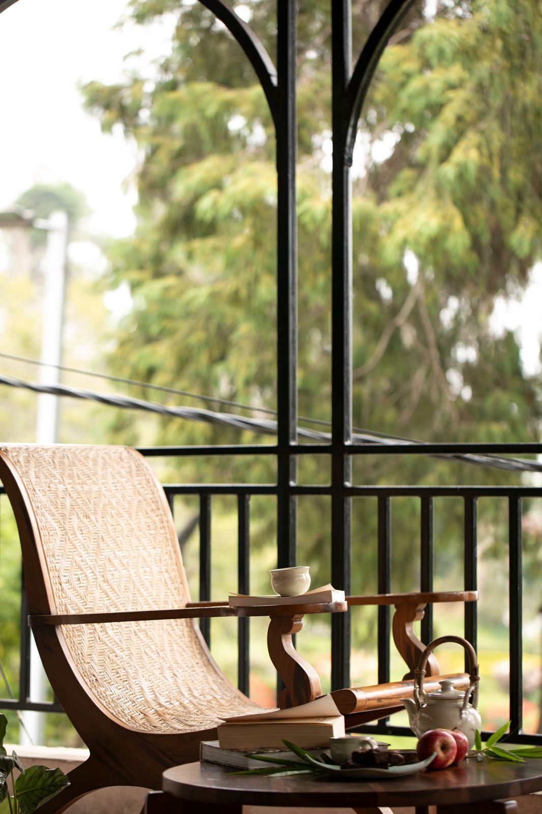 A wooden chair with a woven backrest, a small table with a teapot, a cup, and a book, and a view of trees through a black metal railing.
