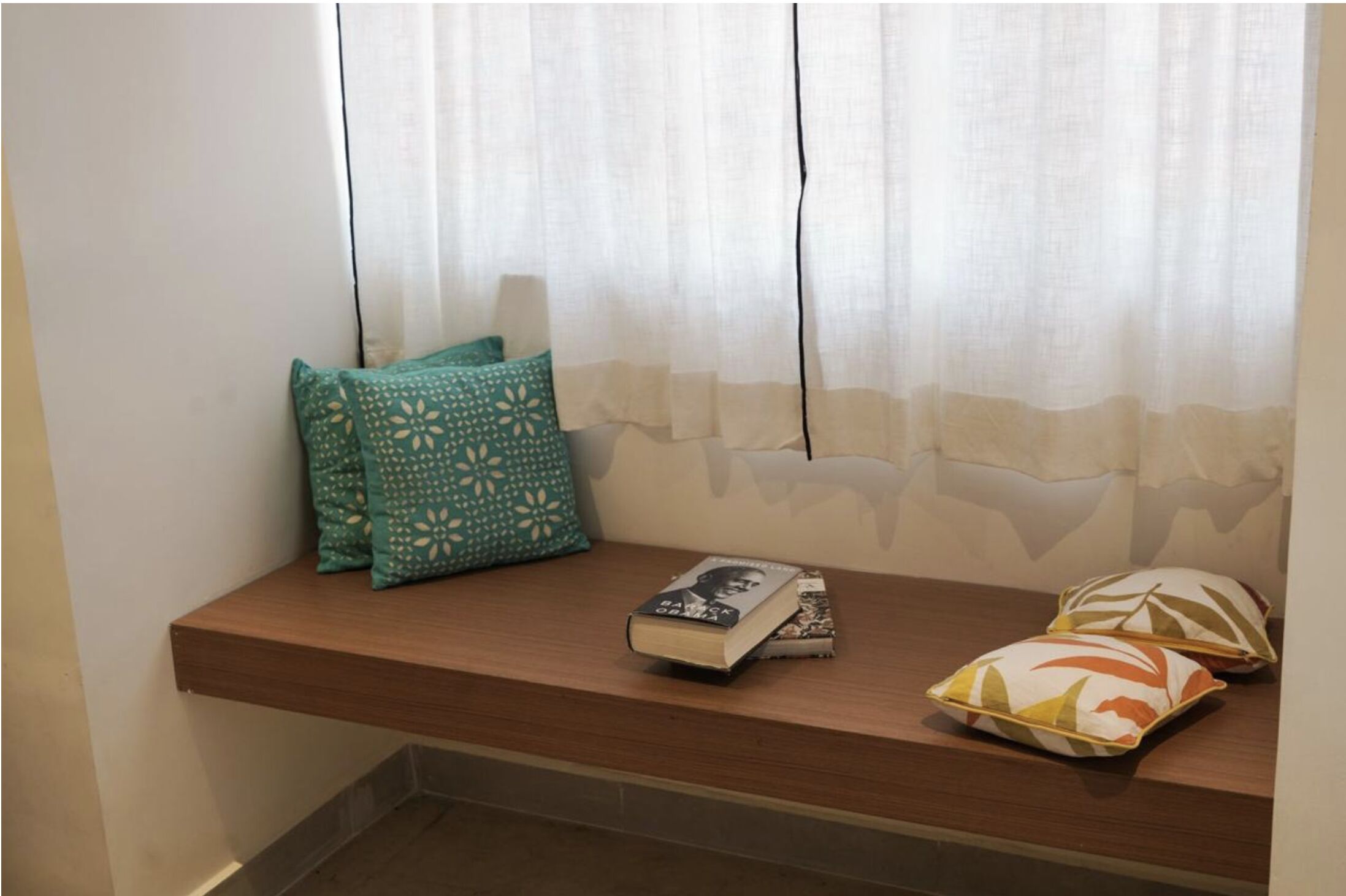A wooden bench with pillows and books against a wall with sheer curtains.
